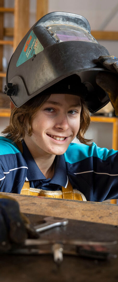 Daily Informer Student Lifts Their Welding Helmet While Working at a Metalwork Bench